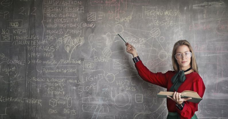 Algebra - Serious female teacher wearing old fashioned dress and eyeglasses standing with book while pointing at chalkboard with schemes and looking at camera